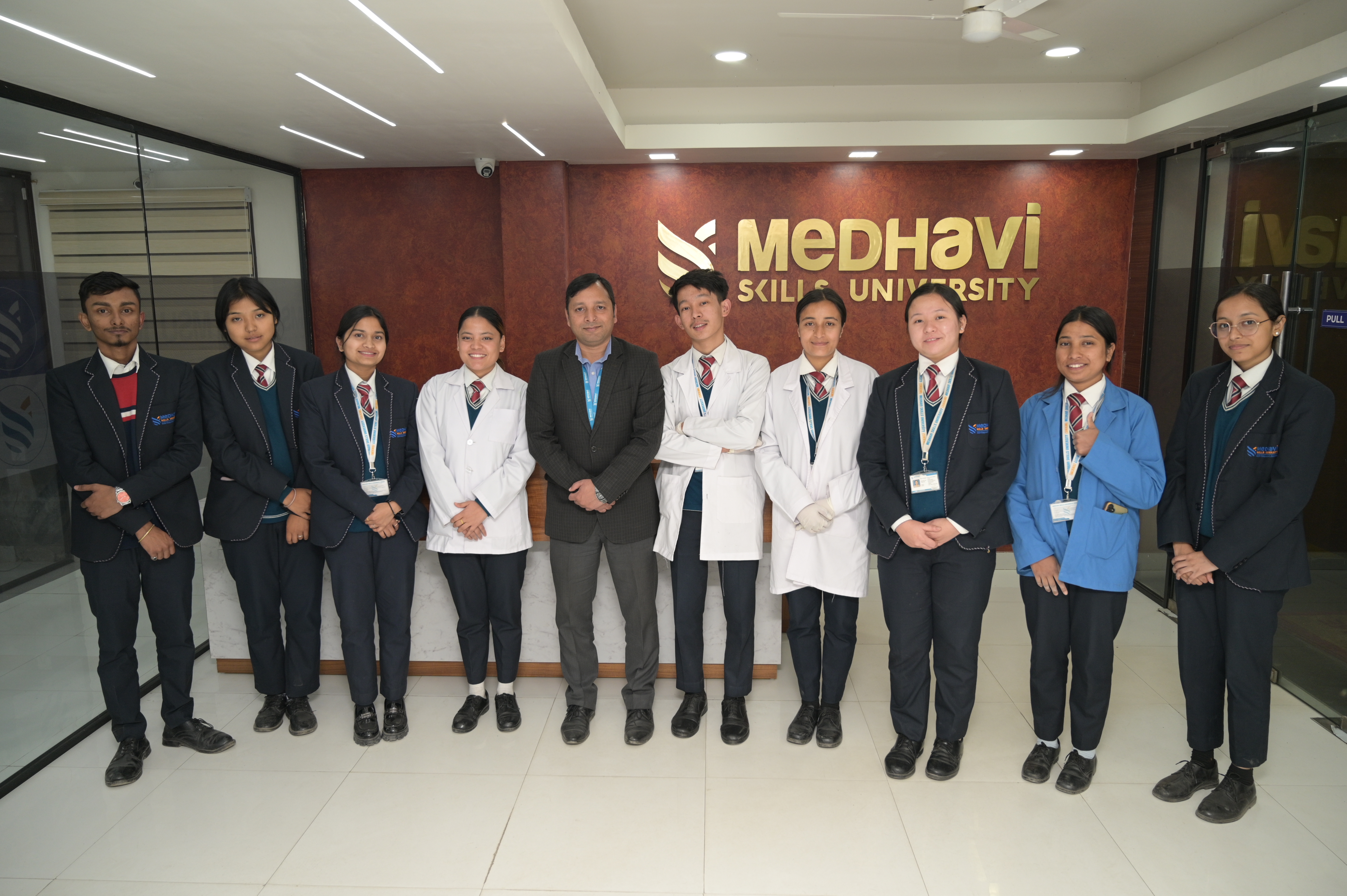 A group of students and a faculty member stand smiling in a row in front of a "Medhavi Skills University" sign. They wear uniforms, some in lab coats.