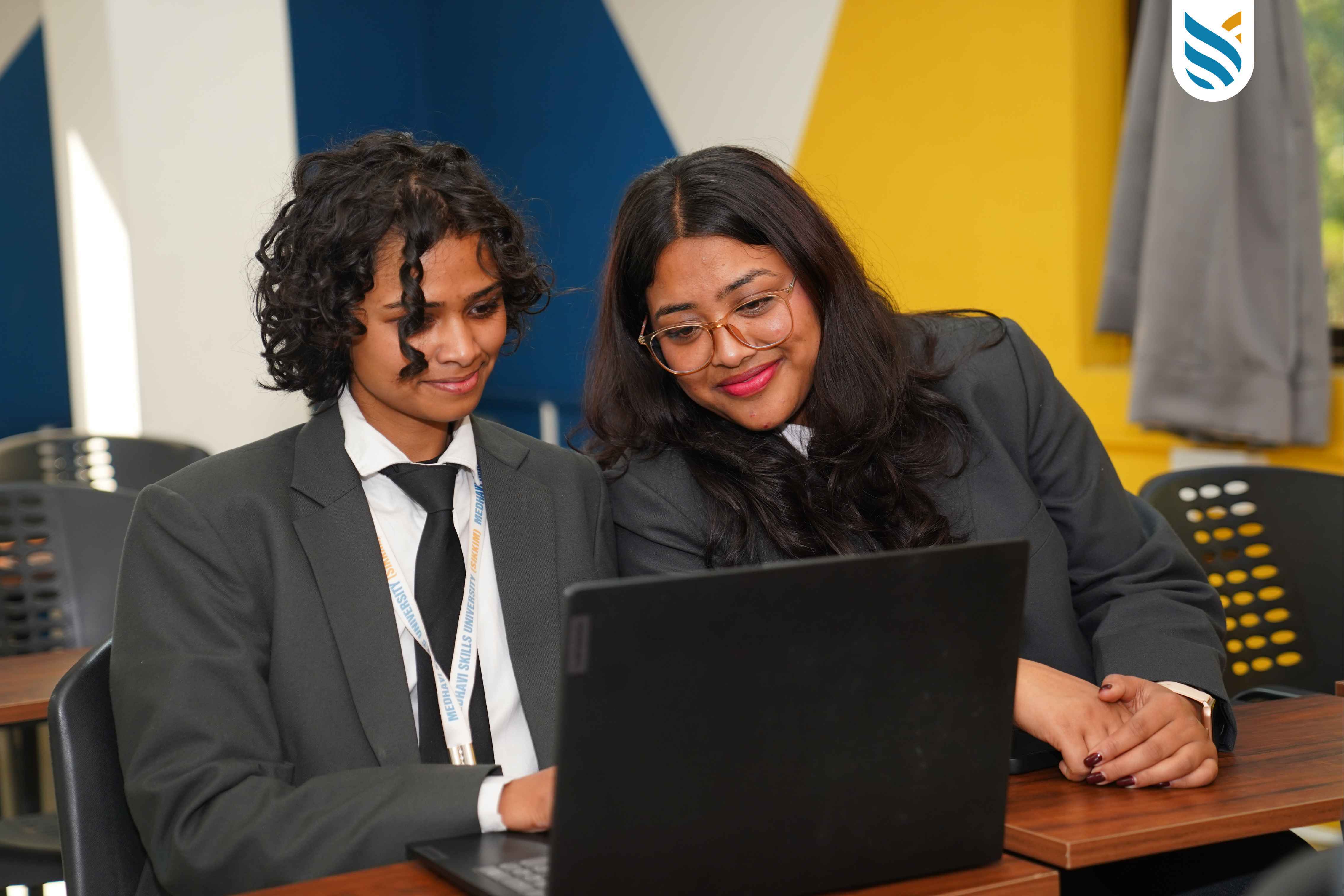 Two people in business attire sit together at a desk, looking at a laptop screen and smiling in a brightly colored classroom.