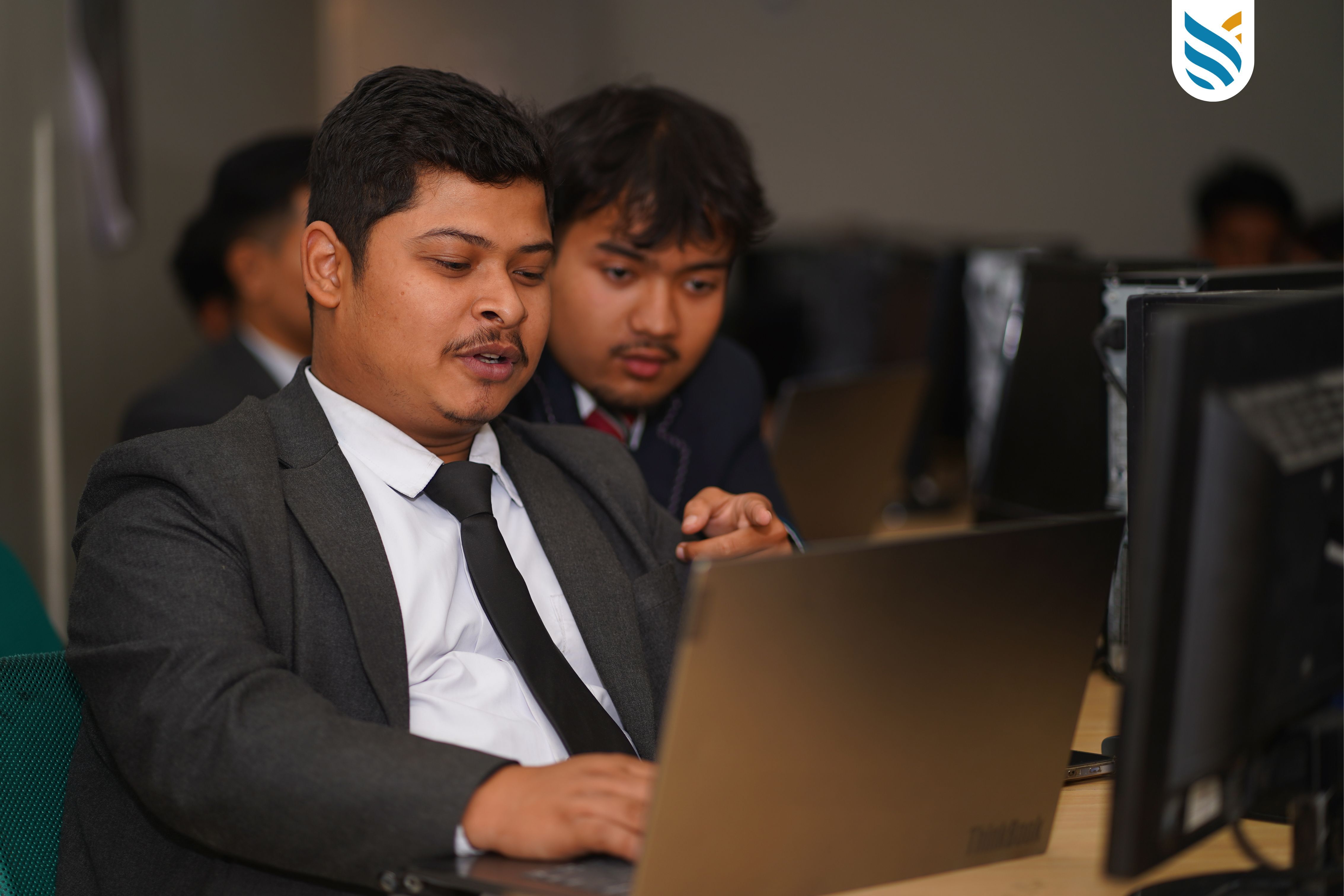 Two men in business attire focus intently on a laptop screen in an office setting, suggesting collaboration or learning. There are computers in the background.