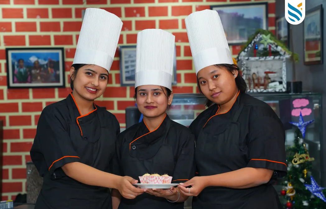 Three chefs in black uniforms and white hats cheerfully hold a dessert plate. They stand against a brick wall, conveying pride and teamwork.