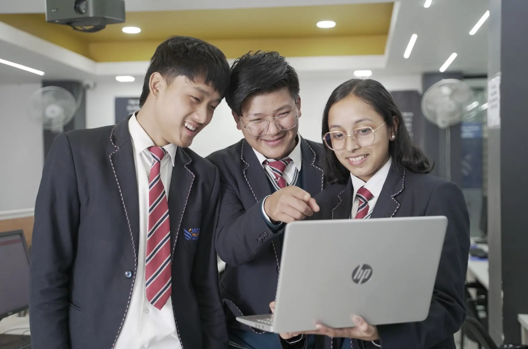 Three students in matching uniforms, smiling and engaged, gather around a laptop in a bright classroom. They emit a sense of teamwork and learning.